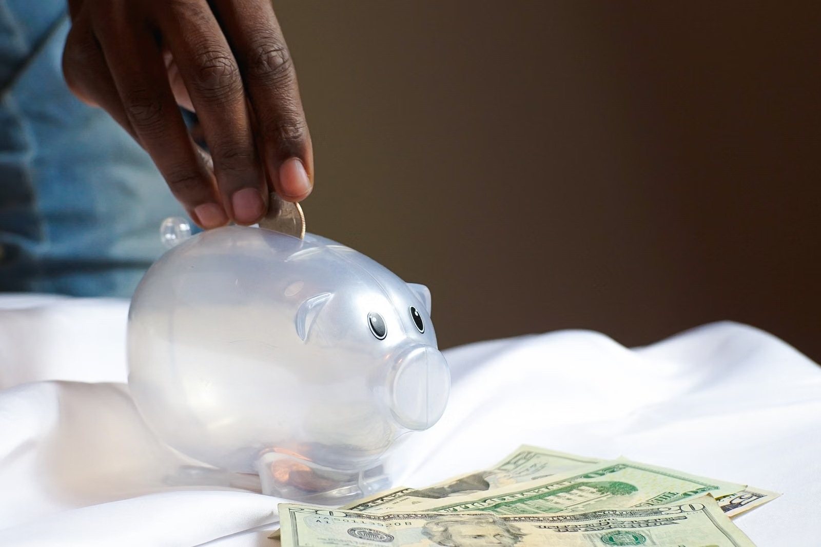 Hand dropping a coin into a clear piggy bank beside US dollar bills